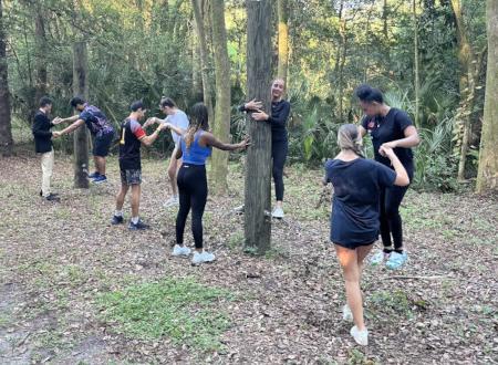 A photo of students on a wood trail holding hands around trees.